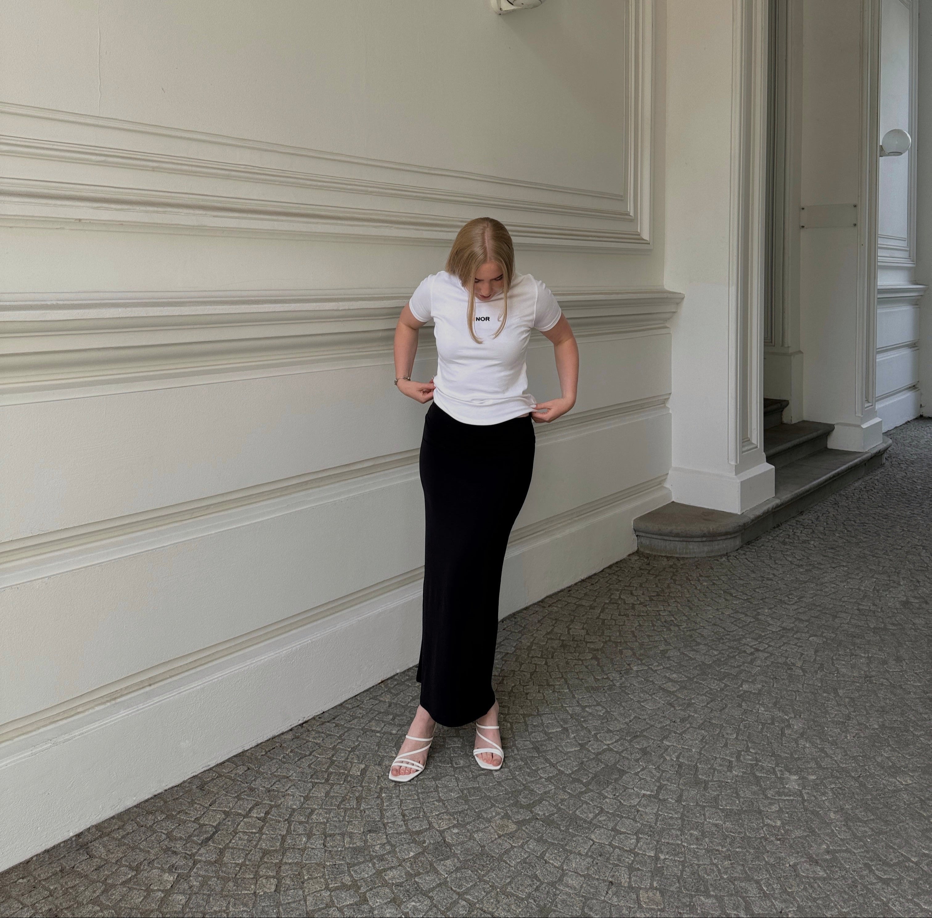 Woman wearing the F1 signature tee styled with a black maxi skirt and white heeled sandals standing in a hallway with a white wall and carpeted floor.