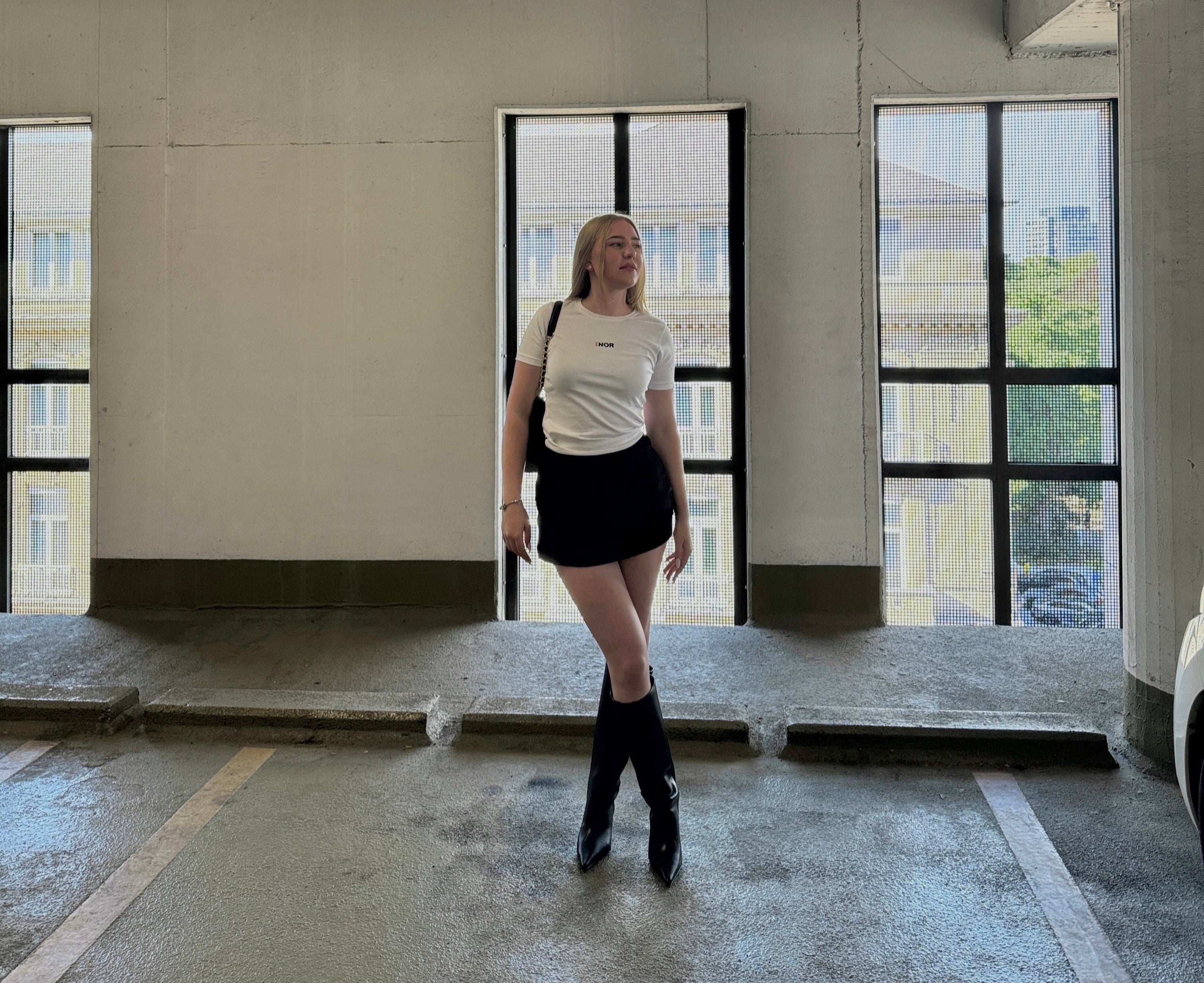 Person wearing the F1 signature tee styled with a short black skirt and knee high heeled boots standing in a large, parking garage with high ceilings and large windows.